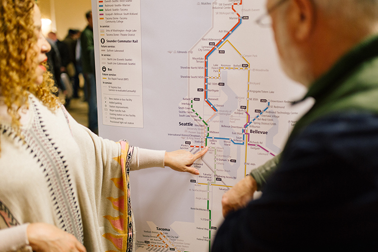 Dos personas de pie frente a un mapa grande del área de Seattle, que presenta las líneas existentes y futuras del servicio de Sound Transit. 